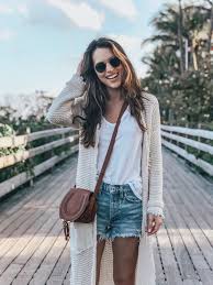 What to Wear in the Bahamas A woman with long brown hair and sunglasses smiles while walking on a wooden bridge. She's wearing a white top, denim shorts, and a beige cardigan.