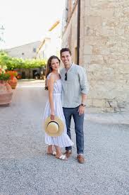 What to Wear in Tuscany A smiling couple poses in a sunny, cobblestone alley. She wears a white dress, holding a hat, and he wears a gray shirt and jeans, exuding a relaxed vibe.