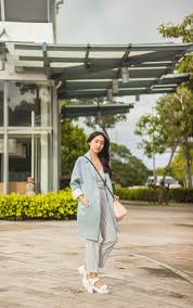 What to Wear in the Philippines A woman in a light blue coat stands outdoors on a paved walkway, surrounded by greenery and modern architecture, exuding a serene and stylish vibe.