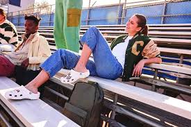đ Women's Crocs: The Ultimate Guide to Comfort, Style, and Fit A woman in a varsity jacket and jeans lounges on stadium bleachers, wearing Crocs. She appears relaxed and content under sunny skies.
