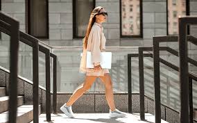 👟 The Ultimate Guide to Women's Casual Sneakers A woman in a tan outfit and white sneakers confidently walks up steps, holding a laptop. She's outdoors, with a modern building in the background.