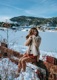 Winter Packing List: What to Pack for a Cold Weather Trip A smiling woman sits on a snow-covered fence, wearing a cozy jacket and red skirt. Snowy mountains and a frozen lake serve as a scenic backdrop.