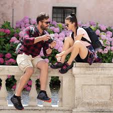 The Ultimate Guide to Travel Shoes for a Round-the-World Trip A man and woman sit on a stone ledge by bright pink flowers. The man shows a camera to the woman. Both wear casual attire and backpacks, smiling.