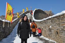 What to Wear to the Great Wall of China A woman in a black coat smiles on a snowy section of the Great Wall of China. Yellow flags line the path, with clear blue skies overhead.