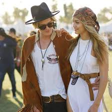 What to Wear to Coachella Music Festival A couple at a festival, dressed in bohemian style. The man wears a hat and jacket; the woman in a white dress and headscarf, both smiling in a sunny outdoor setting.