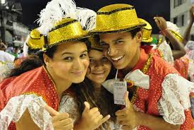 What to Wear to Carnival      in Rio Three people in festive outfits with gold hats and red costumes smile and pose happily. The atmosphere is joyful and celebratory, suggesting a carnival or parade.