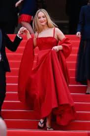 What to Wear to Cannes Film Festival A woman in an elegant red gown walks down a red-carpeted staircase, smiling and holding someone's hand. The scene conveys glamour and sophistication.