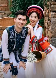 What to Wear in Yunnan, China A smiling couple in traditional clothing poses outdoors. The woman holds a basket of white flowers. Stone wall and greenery in the background convey a cheerful tone.