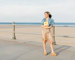 What to Wear in Virginia Beach on Vacation A person stands on a seaside promenade, holding a yellow book, wearing a denim jacket and floral skirt. The background shows a calm beach and ocean.