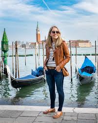 What to Wear in Venice in Spring A woman in sunglasses and a brown jacket stands smiling in front of gondolas on a canal in Venice, with a bell tower in the background under a blue sky.