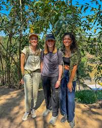 What to Wear in the Amazon Rainforest Three women stand on a dirt path in a lush jungle, smiling. They wear casual hiking attire under a clear blue sky, exuding a cheerful, adventurous mood.